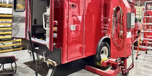 Red fire truck undergoing maintenance inside a service facility, with technicians' equipment, diagnostic tools, and repair systems positioned around the vehicle for fleet maintenance and emergency vehicle servicing.