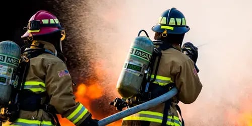 Two firefighters wearing full protective gear and self-contained breathing apparatus (SCBA) work together to control a large fire, spraying water into intense flames during an emergency response operation.