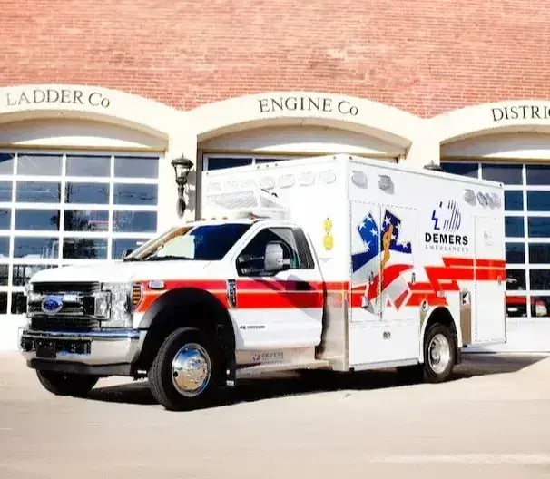 Demers Type I ambulance with a truck-based chassis, modular patient compartment, and exterior storage compartments parked outside a fire station.