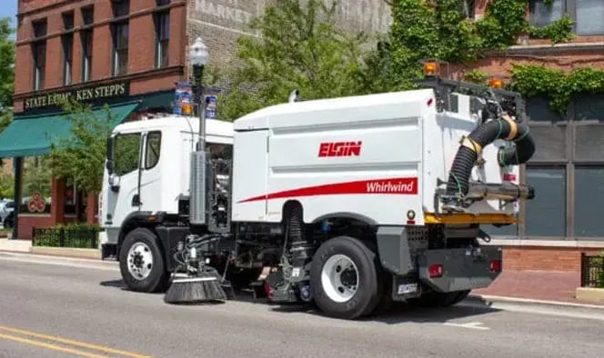 Elgin Whirlwind vacuum street sweeper operating on a downtown city street, featuring powerful vacuum sweeping technology for municipal street cleaning, debris removal, and urban roadway maintenance.