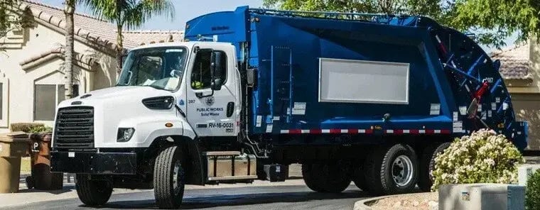 A blue and white Public Works rear-loading garbage truck traveling through a residential neighborhood with palm trees and stucco homes.