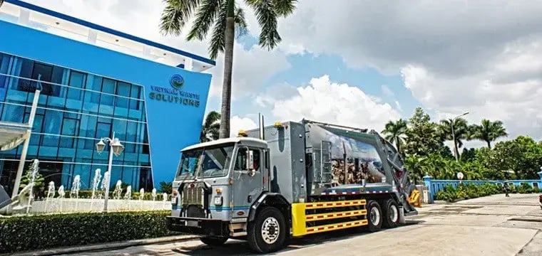 Automated rear loader garbage truck parked outside a modern waste solutions facility with palm trees and landscaping, highlighting commercial waste collection and fleet operations.