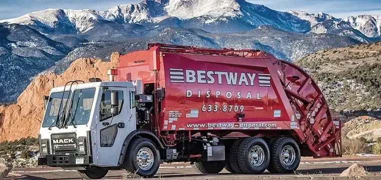 Red Bestway Disposal McNeilus rear loader garbage truck parked on a roadway with snow-capped mountains and rocky terrain in the background, showcasing a residential waste collection vehicle.