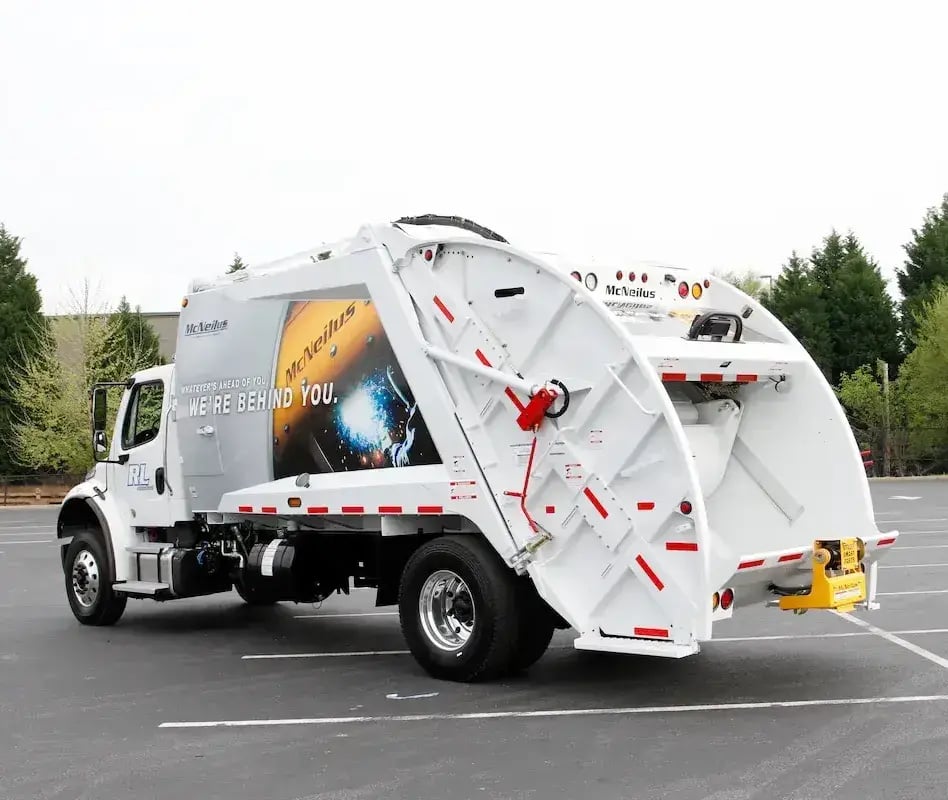 McNeilus rear loader garbage truck parked in a lot, featuring a rear-loading hopper and compaction system used for residential curbside trash collection and municipal solid waste management services.