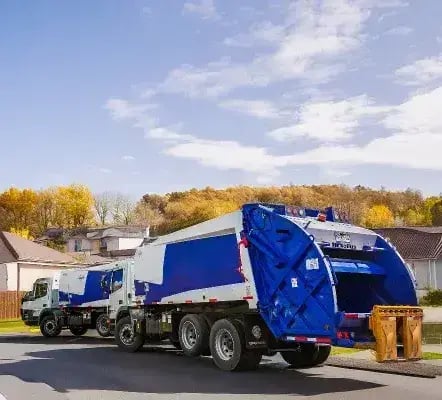 Two McNeilus split-body rear loader trucks driving through a residential neighborhood, designed for simultaneous recycling and waste collection with dual-compartment bodies that improve route efficiency and municipal sanitation operations.