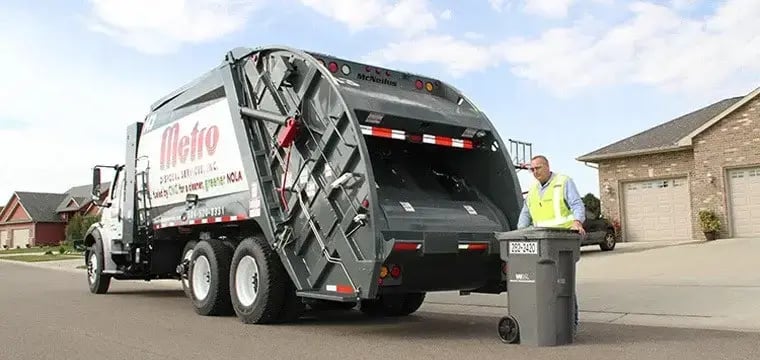 Metro rear loader garbage truck collecting a residential wheeled trash cart at the curb in a suburban neighborhood, with a sanitation worker preparing for waste pickup service.