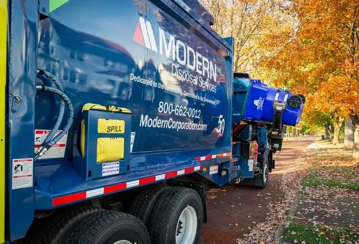 Modern Disposal Services automated side loader truck collecting a blue recycling cart along a residential street lined with autumn trees, demonstrating automated curbside recycling collection.