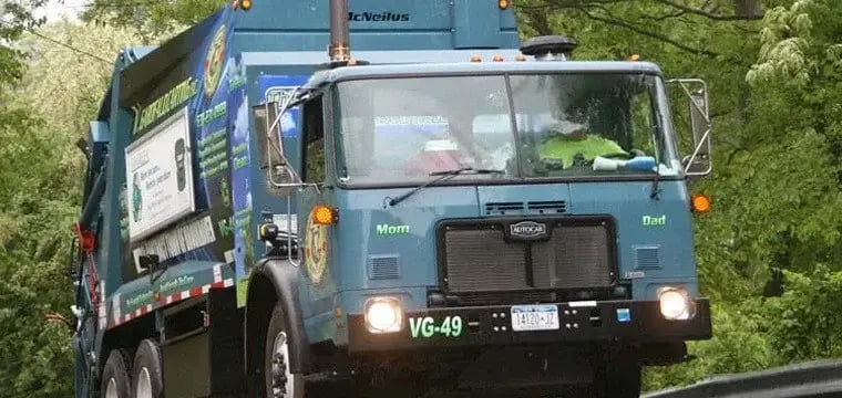 Municipal rear loader garbage truck traveling on a tree-lined roadway, used for residential waste collection and community sanitation services with a high-capacity rear-loading compaction body.