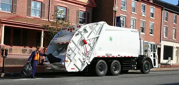 Municipal rear loader garbage truck collecting residential trash bags along a city street lined with historic brick buildings, demonstrating urban curbside waste collection and sanitation services.