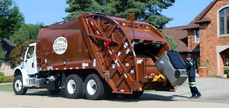 Rear loader garbage truck collecting a residential wheeled trash cart at a curbside pickup location in a suburban neighborhood, operated by a sanitation worker during waste collection service.
