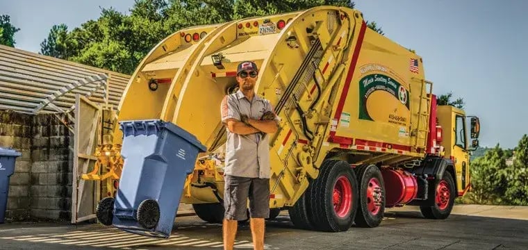 Rear loader garbage truck with an automated cart tipper and sanitation worker standing beside a residential wheeled trash cart, demonstrating efficient curbside waste collection and municipal sanitation services.