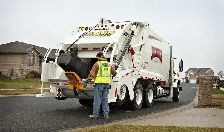Rear loader garbage truck equipped with an automated cart tipper collecting a residential wheeled trash cart, with a sanitation worker assisting during curbside waste collection in a suburban neighborhood.