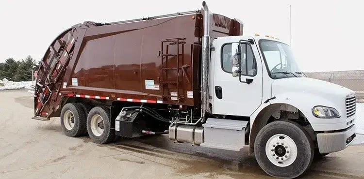 Brown rear loader garbage truck with a white cab parked at a fleet facility, featuring a rear-loading compaction body designed for residential curbside trash collection and municipal solid waste management operations.