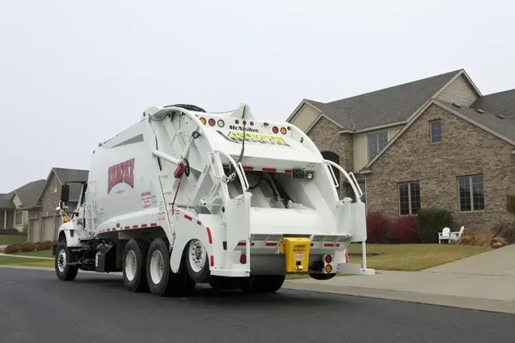 McNeilus rear loader garbage truck driving through a suburban neighborhood, featuring a rear-loading compaction body designed for residential curbside trash collection and municipal solid waste management services.