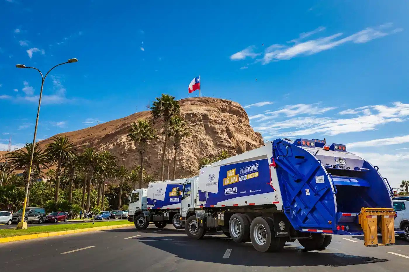 McNeilus rear loader garbage trucks operating in a municipal waste collection fleet near a landmark hillside and large national flag, highlighting urban sanitation and solid waste management services.