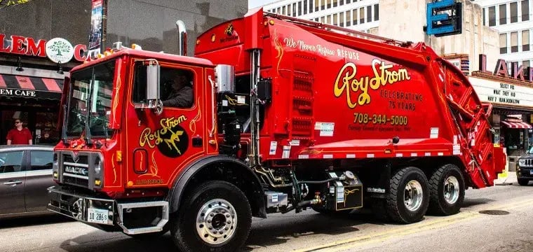 Roy Strom rear loader garbage truck driving through a downtown urban area, providing residential and commercial waste collection services with a high-capacity rear-loading compaction body.