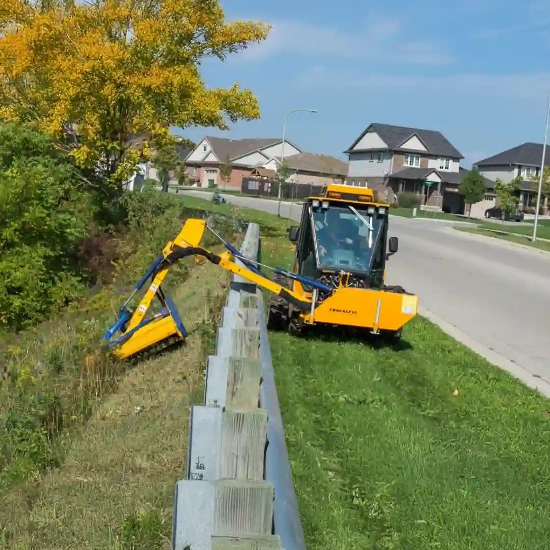 Trackless MT7 municipal tractor equipped with a front-mounted boom mower for trimming grass and vegetation along roadsides, ditches, and guardrails.