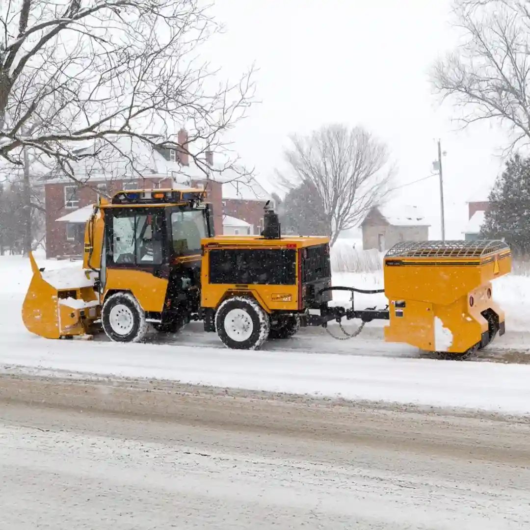 Trackless MT7 compact utility tractor with a front snowplow and tow-behind spreader applying deicing materials during winter maintenance operations.