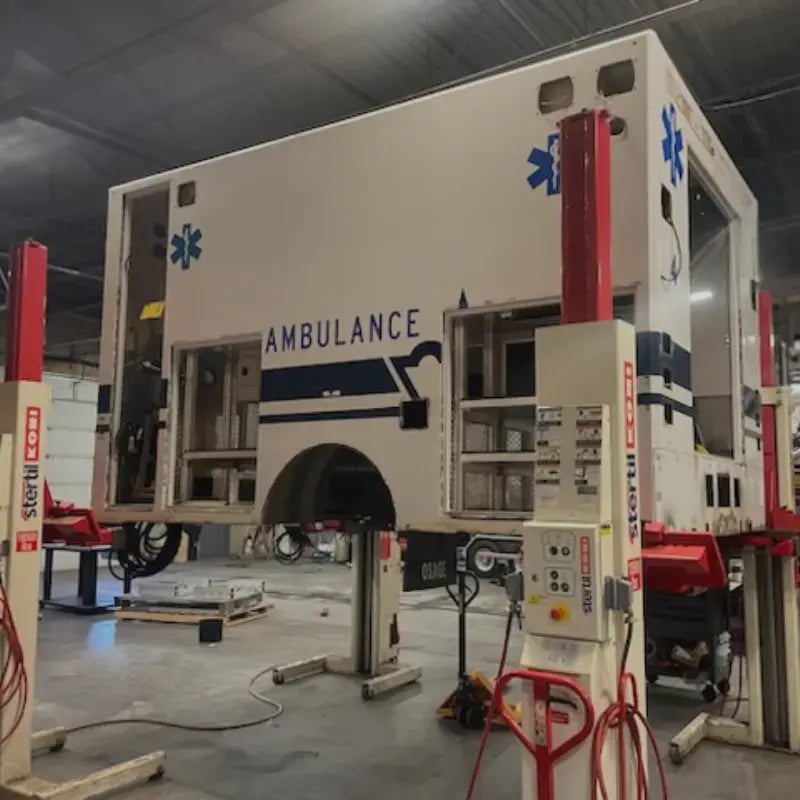 Ambulance patient module elevated on hydraulic lifts during remounting, demonstrating emergency vehicle refurbishment and box transfer services.