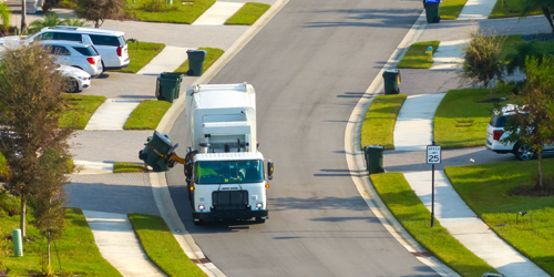 Automated side loader garbage truck collecting a residential wheeled trash cart along a suburban neighborhood street, demonstrating efficient curbside waste collection with a single-operator automated lifting system.
