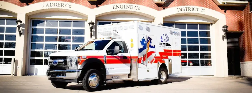 Demers Type I ambulance with a truck-based chassis, modular patient compartment, and exterior storage compartments parked outside a fire station.