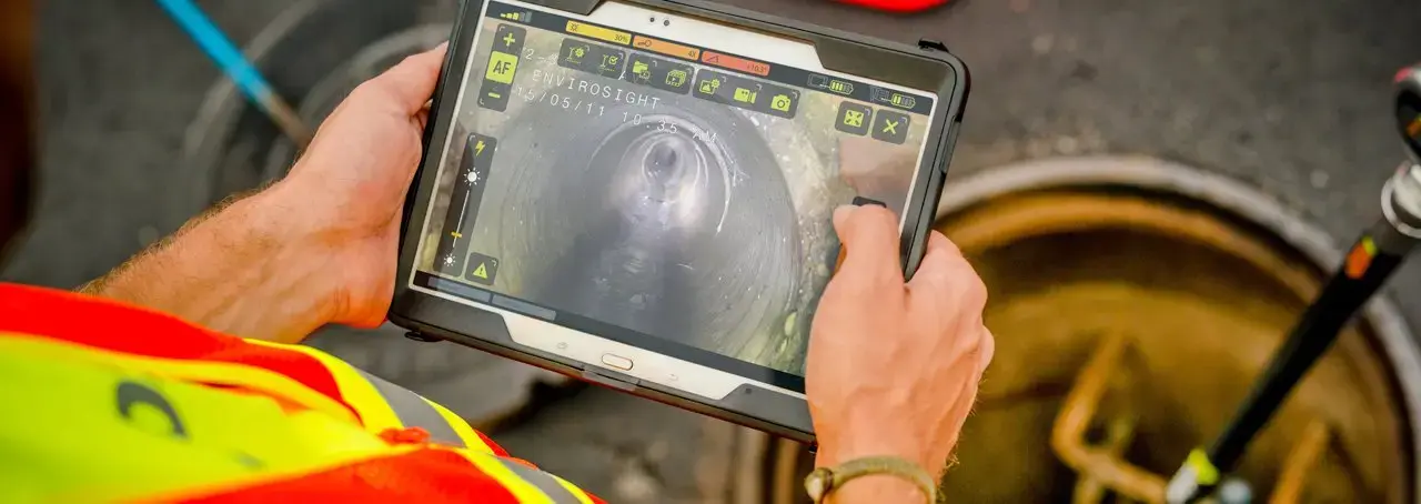 Technician using a tablet to perform a CCTV sewer pipe inspection, viewing live video from a pipeline inspection camera to assess underground sewer conditions, identify defects, and support infrastructure maintenance.