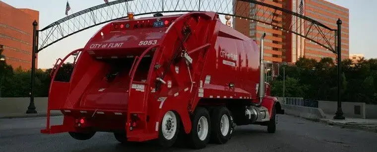 City of Flint rear loader garbage truck traveling through an urban downtown area, providing municipal solid waste collection services with a high-capacity rear-loading compaction body.