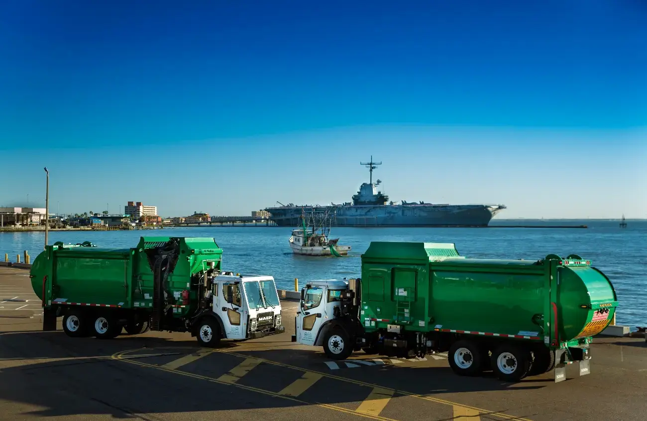 Two rear loader garbage trucks parked along a waterfront roadway, showcasing municipal solid waste collection vehicles operating in a coastal urban environment with a harbor and ship in the background.