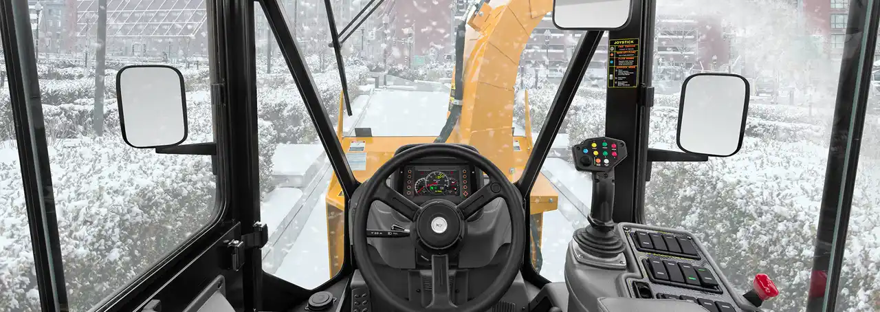 Operator's view from inside a Trackless snow removal vehicle cab showing the steering wheel, controls, and front-mounted snow blower clearing a snow-covered pathway.