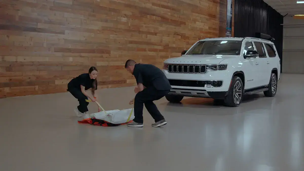 Two individuals deploying a fire suppression blanket beside a vehicle indoors, demonstrating emergency response procedures and fire safety equipment use.