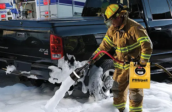 Firefighter operating a foam application system beside a pickup truck, demonstrating fire suppression equipment used for flammable liquid and vehicle fire response.