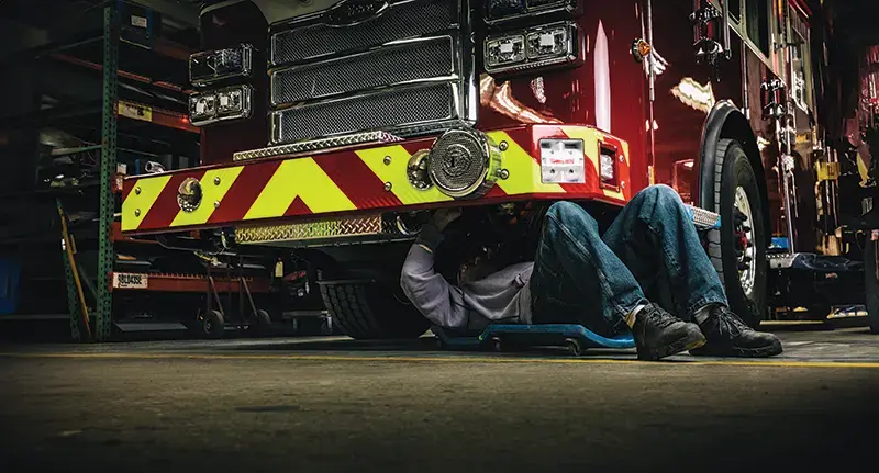 Person working under a Pierce fire truck in a garage, inspecting the front undercarriage and components.